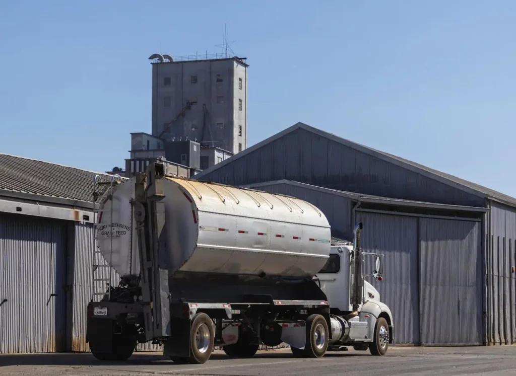 Truck transporting a grain tank parked in front of an industrial warehouse with a tall silo in the background under a clear blue sky.