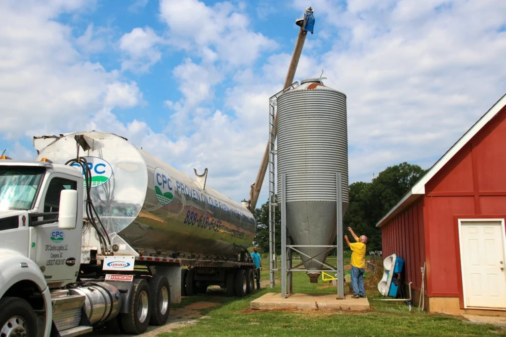 A truck unloading grain at a silo beside a red barn on a farm.